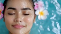 Young Asian woman with glowing hydrated skin relaxing in spa water with tropical plumeria flowers, eyes closed in peaceful wellness moment.