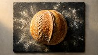 Freshly baked round loaf with scored pattern on dark floured surface, overhead view for bakery, food photography, culinary content and recipe blogs.