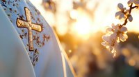 Ornate golden cross on white fabric with soft bokeh sunlight and delicate spring cherry blossoms creating spiritual atmosphere for religious ceremonies and celebrations.