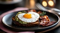 Sunny side up egg on dark toast with mashed avocado and crispy bacon strips on black plate, breakfast setting with warm bokeh lights in background.