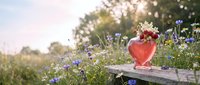 Red heart-shaped vase with wildflowers on wooden fence in garden during golden hour sunset creating romantic atmosphere for Valentine's Day or wedding decor.