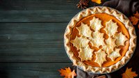 Homemade pumpkin pie decorated with maple leaf pastry cutouts on dark wooden background with autumn leaves and copy space for seasonal menu.