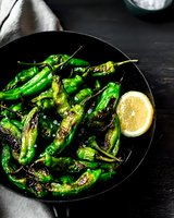 Grilled green shishito peppers served on black plate with fresh lemon slice. Dark moody food photography with dramatic lighting and rustic styling.