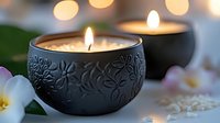 Glowing aromatherapy candles in decorative black ceramic holders with floral pattern, white plumeria flowers and soft bokeh lights creating spa atmosphere.