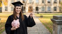 Young Caucasian female graduate in black cap and gown holding pink piggy bank on university campus, celebrating education investment and financial planning.