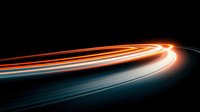 Long exposure light trails on dark background create dynamic motion effect with glowing orange and blue streaks curving along highway at night.