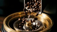 Coffee beans pouring from wooden scoop into vintage brass grinder with warm lighting for cafe and kitchen decor.