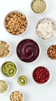 Healthy breakfast bowl ingredients with acai smoothie, granola, nuts, seeds, coconut flakes and fresh kiwi slices arranged on white background.