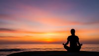 Person meditating in lotus pose on beach at sunset with colorful sky and ocean waves creating peaceful wellness atmosphere for mindfulness practice.