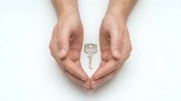 Male hands forming protective heart shape around silver house key on white background symbolizing home security and real estate protection.