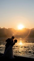 Romantic couple silhouette embracing at golden sunset by lake with lens flare and warm light reflection on water creating intimate wedding moment.