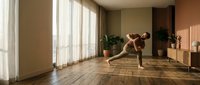 Woman with reddish-brown hair, light skin tone, performing a plank exercise in a bright, minimalist home gym with wooden flooring, potted plants, and natural light from large windows.