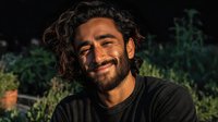 Happy Middle Eastern man with curly hair and beard smiling outdoors in natural garden setting with warm golden hour lighting for lifestyle content.