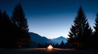 Illuminated camping tent glowing warmly against mountain silhouettes at twilight with evergreen forest creating peaceful outdoor adventure scene for hiking enthusiasts.