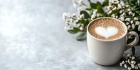Hot cappuccino with heart-shaped milk foam art served in white ceramic cup alongside fresh baby breath flowers on light gray concrete background with copy space.