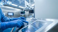 Surgeon in blue scrubs and gloves operating a touchscreen control panel in a modern sterile operating room environment.