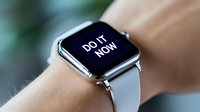 Modern smartwatch displaying motivational message "Do it now" on black screen with white text, worn on wrist with light leather band against blurred background.