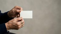 Senior man with weathered hands holding blank white business card against neutral gray background, suitable for corporate identity, professional services, and business marketing materials.