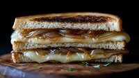 Grilled cheese sandwich with melted cheese and caramelized onions on toasted bread, close-up view showing layers of gooey cheese against dark background.