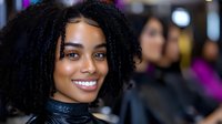 Young African American woman with natural curly hair and bright smile wearing black outfit against blurred background in indoor setting.