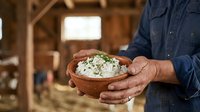 Farmer holding bowl of fresh cheese curds in rustic barn setting, showcasing artisanal dairy production and farm-to-table food preparation.