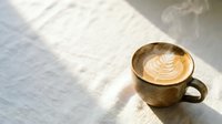 Steaming hot latte with leaf pattern foam art in ceramic cup on white wooden table with natural sunlight shadows creating cozy morning coffee shop atmosphere.