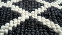 Handwoven wool rug with geometric diamond pattern in charcoal gray and cream colors. Traditional textile craft showcasing intricate knitting technique and texture.