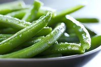Fresh steamed green snap beans with water droplets on dark plate, macro food photography showing texture and glossy surface in natural lighting.