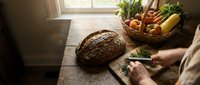 Rustic kitchen scene with fresh bread, vegetables and herbs on wooden table near window. Cozy cooking preparation with natural lighting for healthy lifestyle.