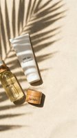 SPF 50 sunscreen, after sun oil, and lip balm on sandy beach with palm shadow, summer skincare essentials flatlay.