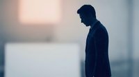 Young adult male in dark business attire standing in contemplative pose against blurred urban background. Suitable for corporate, mental health, depression awareness, and professional lifestyle concepts.