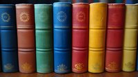 Row of vintage hardcover books with colorful spines in blue, red, yellow and green standing vertically on shelf, showing classic binding and gold decorative details.