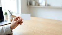 Person holding blank white card at wooden desk with laptop and office supplies in bright workspace.