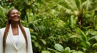 Young woman with braided hair and dark skin tone wearing white blazer smiling outdoors surrounded by lush green foliage and tropical plants.
