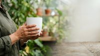 Person holding white cup with blurred green plants in background, suitable for beverage, lifestyle, and wellness marketing materials.