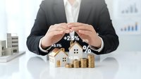 Real estate investor in suit protecting house models with coin stacks and growth percentage signs, symbolizing property investment returns.