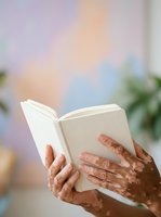 Adult with medium-deep skin tone holding open cream-colored book against soft pastel background with blurred foliage. Suitable for education, literacy, reading, and learning concepts.