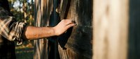Male hand touching wooden fence post outdoors, wearing plaid shirt. Rustic countryside scene for editorial and lifestyle use.