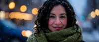 Young Hispanic woman with curly hair wearing green knit scarf smiling warmly during snowfall with bokeh city lights background for winter lifestyle content.