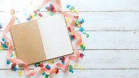 Open notebook with blank pages surrounded by colorful confetti and striped ribbons on white wooden background for celebration planning and party design.