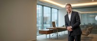 Confident middle-aged Caucasian businessman in dark suit standing in modern office boardroom with large windows and contemporary furniture.