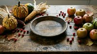 Autumn harvest arrangement with decorative gourds, fresh apples, corn, and empty rustic plate on weathered wooden surface for seasonal styling.