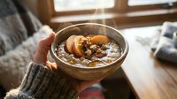 Hands holding warm bowl of oatmeal porridge with apple slices and nuts, cozy breakfast scene with natural window light for food photography and wellness content.