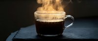 Steaming hot black coffee in a clear glass cup on a dark slate surface, with dramatic moody lighting and rising vapor.