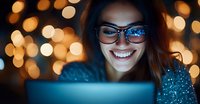 Young woman in glasses smiling while looking at laptop screen at night, with warm bokeh lights in background creating cozy atmosphere. Candid moment captures joy.