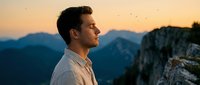 Man with dark hair and light complexion gazing at mountain landscape during golden hour sunset, standing on cliff edge overlooking valleys and peaks, contemplative mood for travel and adventure themes.