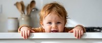 Adorable toddler with curly blonde hair peeking over kitchen counter edge, curious expression, home cooking environment with wooden utensils in background.