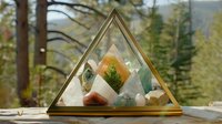 Glass triangular terrarium with healing crystals, minerals and small plants arranged in decorative composition against blurred forest background, creating zen atmosphere.
