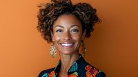 Young African American woman with natural curly hair wearing colorful floral print blouse smiling confidently against orange background, professional studio portrait.