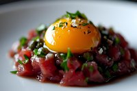 Fresh tuna tartare topped with raw egg yolk, chopped herbs, and sea salt flakes served on white plate, close up view of gourmet appetizer preparation.